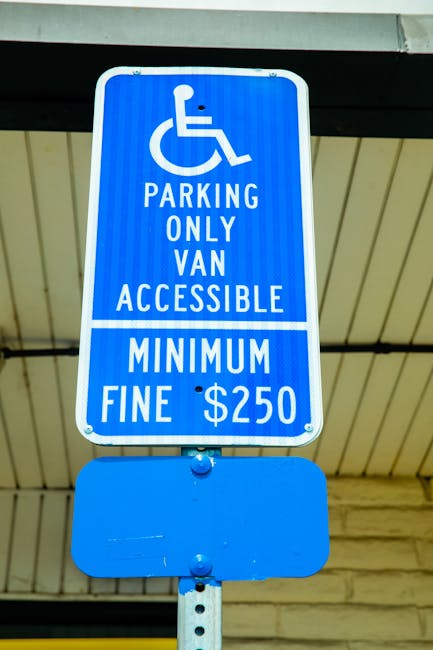 Close-up of a blue parking sign indicating designated parking for disabled individuals and vans, situated outdoors under a roof structure with visible support beams. The sign displays the wheelchair symbol at the top, followed by the text 'PARKING ONLY VAN ACCESSIBLE' and a minimum fine of '250 dollars'. The sign is mounted on a metal pole with a small blank blue panel below it. The environment suggests a loading or unloading area, possibly near a residential or commercial property on Colham Road in Yiewsley, relevant to house removals and moving services offered by Man with Van Yiewsley. The image emphasizes the importance of parking regulations for efficient furniture transport and home relocation logistics.