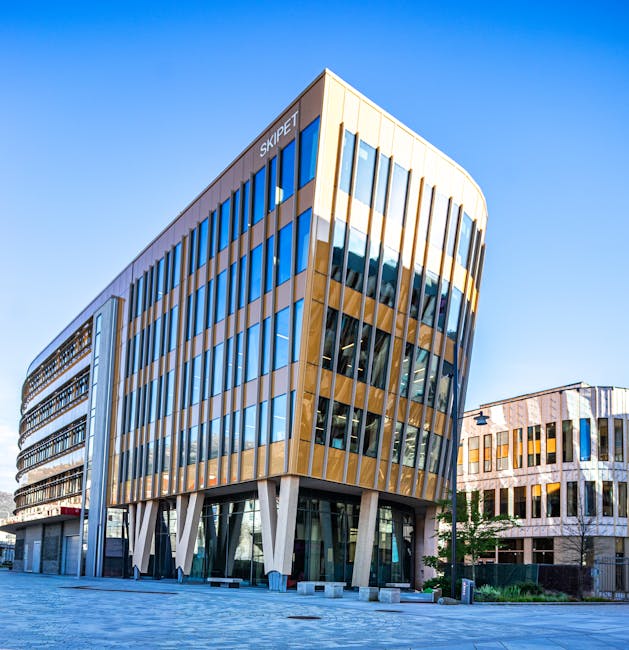 A modern multi-storey office building with a rounded corner located on a paved street, featuring extensive blue-tinted glass windows and golden-brown vertical cladding. The building's ground floor has