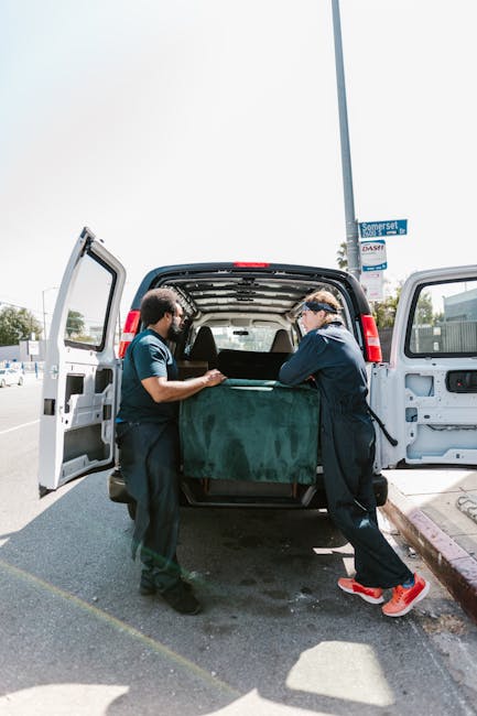 Two movers from Man with Van Yiewsley are engaged in a home relocation task, standing on a pavement next to an open van with its rear doors wide open. One worker, wearing a navy blue uniform, is holding the edge of a large, dark green upholstered piece of furniture wrapped in protective fabric, while the other, dressed in a dark blue shirt and black trousers, is assisting in lifting or stabilising the item. Inside the van, several other boxes and packing materials are visible, suggesting the ongoing process of packing and loading during furniture transport. The van is positioned on a street, near a lamppost which displays a council permit or parking notice, indicating compliance with local regulations for loading activities. Bright daylight illuminates the scene, with a partly cloudy sky overhead, and the background shows a typical urban setting with signs, trees, and other parked vehicles, supporting the context of a professional moving and removal service in Yiewsley.