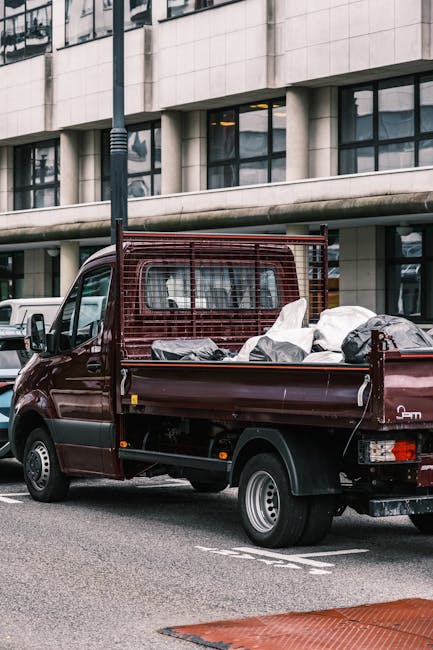 A dark red van parked on a city street in front of a modern building with large glass windows and concrete cladding. The van's open flatbed is loaded with various household items, including cardboard boxes and wrapped furniture, some covered with white protective fabric or plastic. The items are arranged for a home relocation, with the vehicle positioned near the pavement, ready for loading or unloading. A black metal grid extends from the van's side, supporting the cargo during transport. In the background, a white van is partially visible, and a streetlamp is situated along the roadside. The scene is lit by natural light, emphasizing the details of the packed household goods and the urban environment associated with professional removals and moving services, such as those offered by Man with Van Yiewsley.