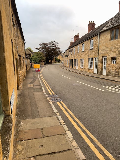 Photograph of Yiewsley High Street showing a narrow residential street with old stone and brick houses lining the curved roadway. The pavement on the left side has various concrete and stone slabs, some of which are uneven or cracked. Two yellow double yellow lines run parallel along the edge of the road. In the background, there is a tree with sparse foliage and a yellow traffic sign on the pavement near orange safety cones. The scene appears overcast with grey skies, and no vehicles or people are visible in the image. This setting illustrates typical surroundings where home relocation or furniture transport might be carried out, highlighting the challenges of narrow access streets for removals services like those provided by Man with Van Yiewsley.