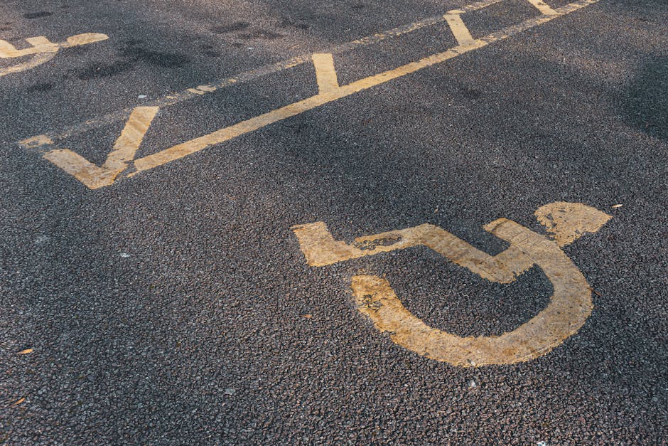 Close-up view of a parking space designated for disabled individuals on Yiewsley High Street, with a worn yellow wheelchair symbol painted on dark asphalt. The parking markings are weathered, with visible cracks and patches. The surface is textured with small stones and gravel embedded in the asphalt. In the background, faint outlines of additional parking space lines can be seen, indicating this area is part of a public parking lot or street parking zone. The scene captures the outdoor environment during daylight, with natural light illuminating the textured surface and faded markings. This image reflects the area where home relocation or moving services by Man with Van Yiewsley might involve accessing or loading items from a nearby property situated on Yiewsley High Street, with considerations for parking and vehicle access during furniture transport or packing and moving processes.
