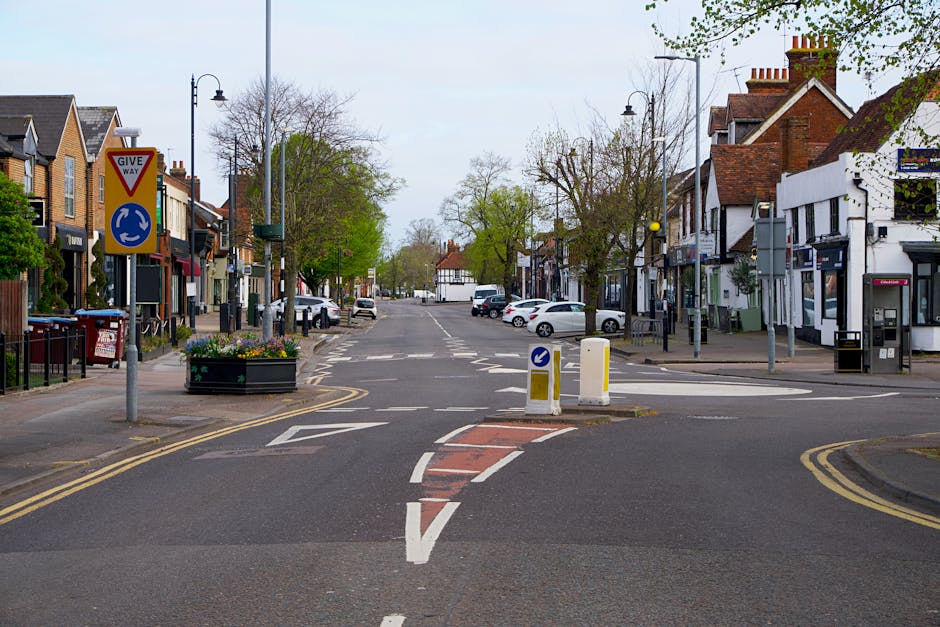 Photograph of Yiewsley High Street showing a narrow residential street with old stone and brick houses lining the curved roadway. The pavement on the left side has various concrete and stone slabs, some of which are uneven or cracked. Two yellow double yellow lines run parallel along the edge of the road. In the background, there is a tree with sparse foliage and a yellow traffic sign on the pavement near orange safety cones. The scene appears overcast with grey skies, and no vehicles or people are visible in the image. This setting illustrates typical surroundings where home relocation or furniture transport might be carried out, highlighting the challenges of narrow access streets for removals services like those provided by Man with Van Yiewsley.