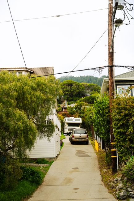 A narrow concrete driveway in Horton Road, Yiewsley, with residential houses on either side, surrounded by greenery and trees. A utility pole with electrical wires runs along the right side of the driveway, with visible transformers and insulators. A metal car parked near the top of the driveway, close to the entrance, with a small caravan positioned behind it. The driveway appears to be used for home relocation or furniture transport, with the surroundings indicating a quiet residential area consistent with house removals in Yiewsley. The scene is well-lit, capturing the typical environment seen during packing and loading processes associated with moving services provided by Man with Van Yiewsley.