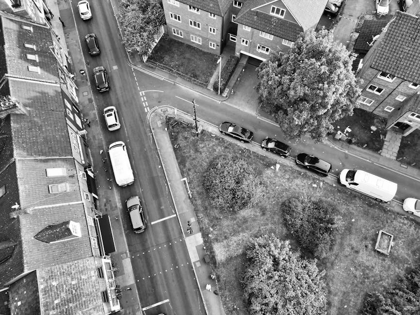 An aerial view showing a narrow residential driveway on Horton Road, Yiewsley, with several vehicles parked along the street and within the driveway, including a white van positioned near the entrance of a property. The scene includes nearby houses with brick facades, windows, and pitched roofs, as well as trees and a small grassy area with bushes and landscaping. A moving company, Man with Van Yiewsley, is visible organizing the loading process, with furniture, cardboard boxes, and packing materials near the van, suggesting a home relocation or furniture transport operation. The area is well-lit with natural daylight, capturing the logistics of house removals in a tight urban setting, emphasizing careful loading and parking arrangements relevant to professional moving services.