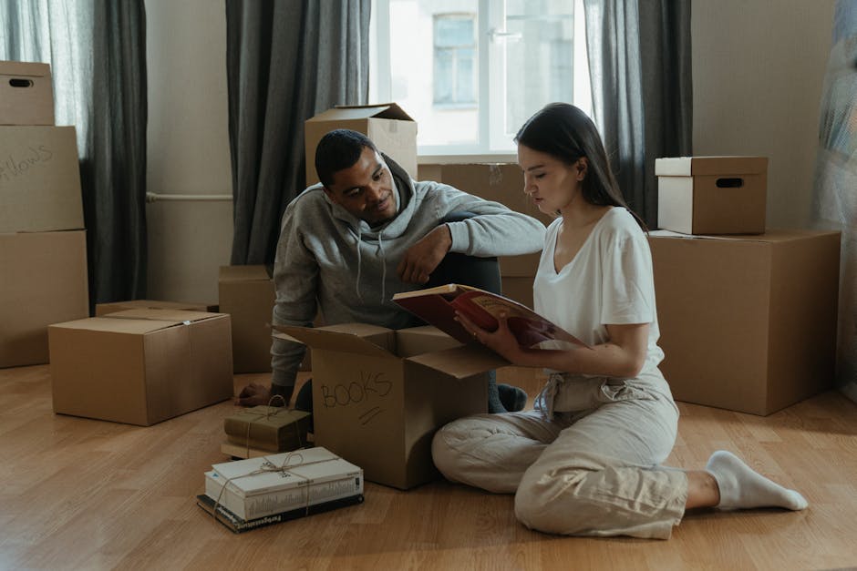 A man and woman are inside a room with light wooden flooring, surrounded by numerous cardboard boxes, some labeled with contents such as 'books,' and others plain. The woman is seated on the floor, wearing light-colored pajamas, and is holding an open book or folder. The man, dressed in a gray hoodie and dark pants, is kneeling beside her, leaning forward with one arm resting on a box, observing the contents. Several boxes are stacked against the wall near a window with curtains, and some are on the floor, ready for packing or transport. The scene captures a home relocation or packing and moving process, with the boxes likely prepared for a house removal. Man with Van Yiewsley, a professional removals company, often handles similar packing and furniture transport tasks, as suggested by this interior setting staged for moving activities, including the careful handling of boxed items in a typical domestic environment.