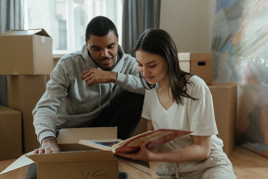 Two individuals, a man and a woman, are engaged in loading or unpacking moving boxes inside a residential property, with several cardboard boxes stacked in the background near a window. The man, dressed in a light gray hoodie, is leaning forward with one hand resting on an open box, while the woman, wearing a white t-shirt, is sitting on the floor holding a book or magazine. The scene captures the packing and moving process, as they prepare belongings for home relocation, with the setting illuminated by natural daylight coming through the window. The workspace includes various cardboard boxes, some open with contents visible, and furnishings suggesting an organized move. Professional removals services like those offered by Man with Van Yiewsley support these activities, providing efficient furniture transport and packing assistance during house moves.