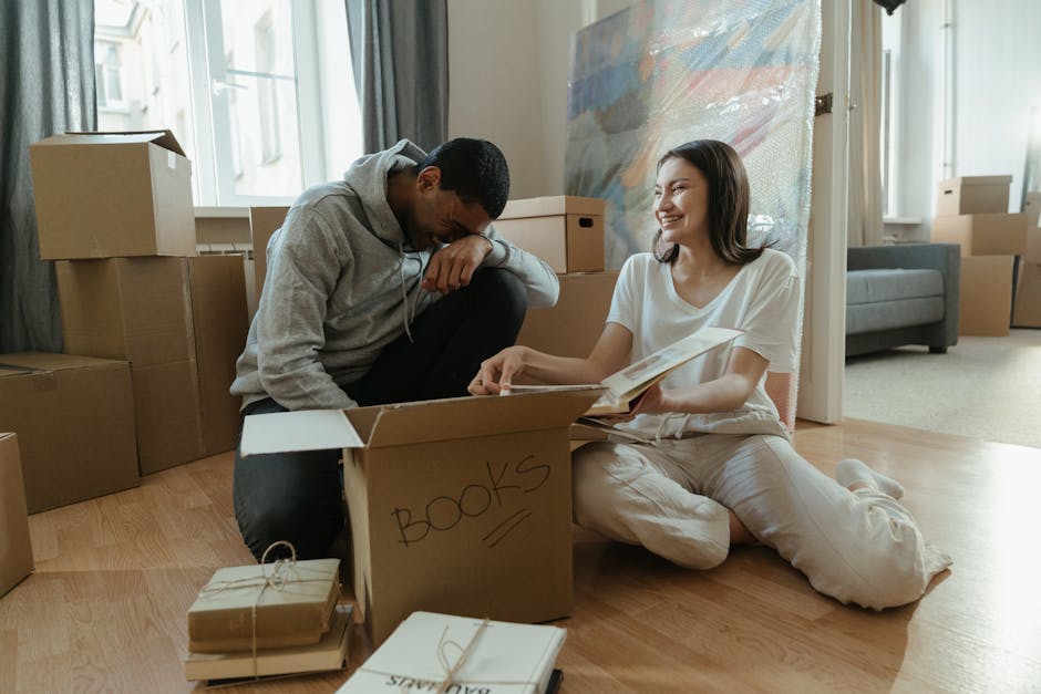 A man and woman are inside a room with light wooden flooring, surrounded by numerous cardboard boxes, some labeled with contents such as 'books,' and others plain. The woman is seated on the floor, wearing light-colored pajamas, and is holding an open book or folder. The man, dressed in a gray hoodie and dark pants, is kneeling beside her, leaning forward with one arm resting on a box, observing the contents. Several boxes are stacked against the wall near a window with curtains, and some are on the floor, ready for packing or transport. The scene captures a home relocation or packing and moving process, with the boxes likely prepared for a house removal. Man with Van Yiewsley, a professional removals company, often handles similar packing and furniture transport tasks, as suggested by this interior setting staged for moving activities, including the careful handling of boxed items in a typical domestic environment.
