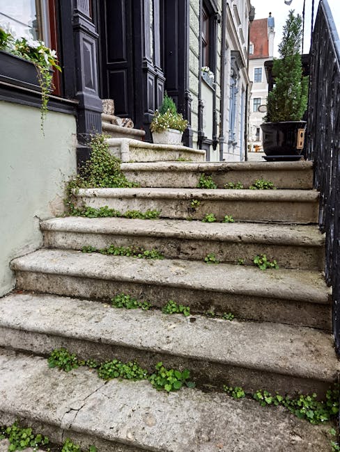 A set of uneven outdoor stone steps leading up to a residential property in Yiewsley. The stairs are constructed from large, weathered stone slabs with visible cracks and erosion, and small green weeds are growing in the gaps between the steps. On either side of the staircase, there are black metal railings and a white wall on the left, with a black rectangular planter containing a small green shrub placed on top of the wall. The property features black painted wooden door and window frames, with potted plants on the windowsills, and the entrance appears narrow, with limited space for large items. The surrounding environment includes neighboring buildings and a paved path at the top of the stairs, suggesting a typical urban residential area. The scene is well-lit with natural daylight, and the image captures the challenges of moving bulky furniture or boxes up or down stairs when access is limited, illustrating the importance of professional removals services such as those provided by Man with Van Yiewsley.