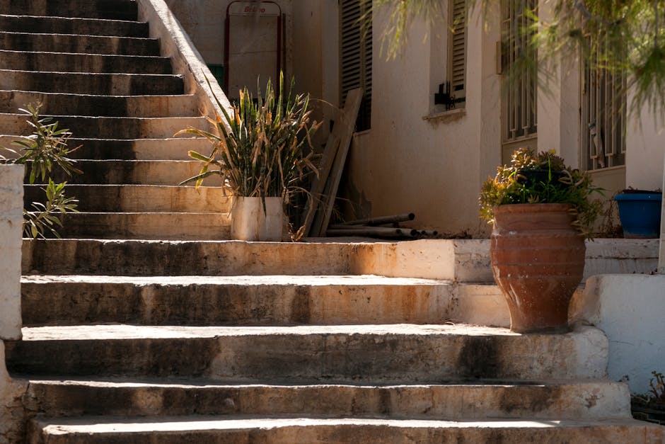 A set of outdoor wooden stairs with several steps leading uphill amid a natural environment, surrounded by dense green foliage, fern leaves, and small plants. The stairs appear weathered and are partially covered by dirt and vegetation, with some steps showing signs of moss growth. To the left, a metal handrail is partially visible, supporting the pathway through the wooded area. This image depicts a challenging access point that may require careful planning for home relocation or furniture transport, especially when moving bulky items. The natural lighting suggests an overcast day or shaded setting, highlighting the texture and details of the greenery and wooden steps. Man with Van Yiewsley’s services would typically address such terrain constraints during house removals involving properties with uneven or difficult access routes, ensuring safe and efficient loading and unloading processes in relocation tasks.