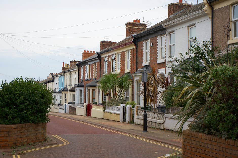 A row of terraced homes in Yiewsley with a paved street in front, showing houses with brick and painted facades, each with small front gardens enclosed by low walls or fences. Some houses feature chimneys, and a few have decorative elements like window trims or small porch areas. The street is lined with greenery, including a large shrub in a brick planter on the left and various plants and palms in front gardens. Utility poles and wires run along the street, and an overcast sky casts diffuse lighting over the scene. This residential area is typical for urban home relocations and furniture transport needs, where careful packing, loading, and transportation by Man with Van Yiewsley are essential for safe moving services. The setting suggests a quiet neighborhood suitable for house removals and moving logistics, with the street background indicative of a typical UK residential street used for professional relocation services.