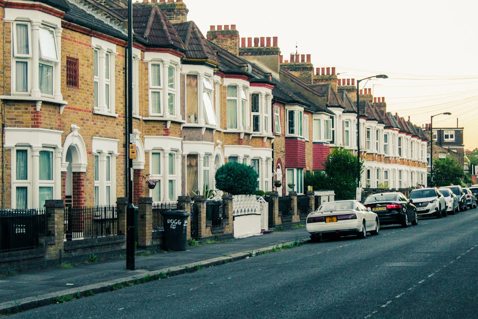 A narrow residential street in Yiewsley lined with brick terraced homes featuring small front porches and gabled roofs. The street is paved with asphalt, with a concrete curb on one side. On the right, a tall wooden fence separates the properties from the pavement, which is partly shaded by nearby trees and bushes. In the distance, several black wheelie bins are visible along the curb, suggesting ongoing home relocation or packing activities. The scene appears to be part of a house removals process, possibly involving furniture transport and loading. The lighting indicates late afternoon or early evening, with shadows cast across the street. The overall environment is calm, with no movement or crowds visible, aligning with a typical moving day within a suburban area, and occasional use of equipment such as trolleys or packing materials by Man with Van Yiewsley for inside packing and outside loading operations.