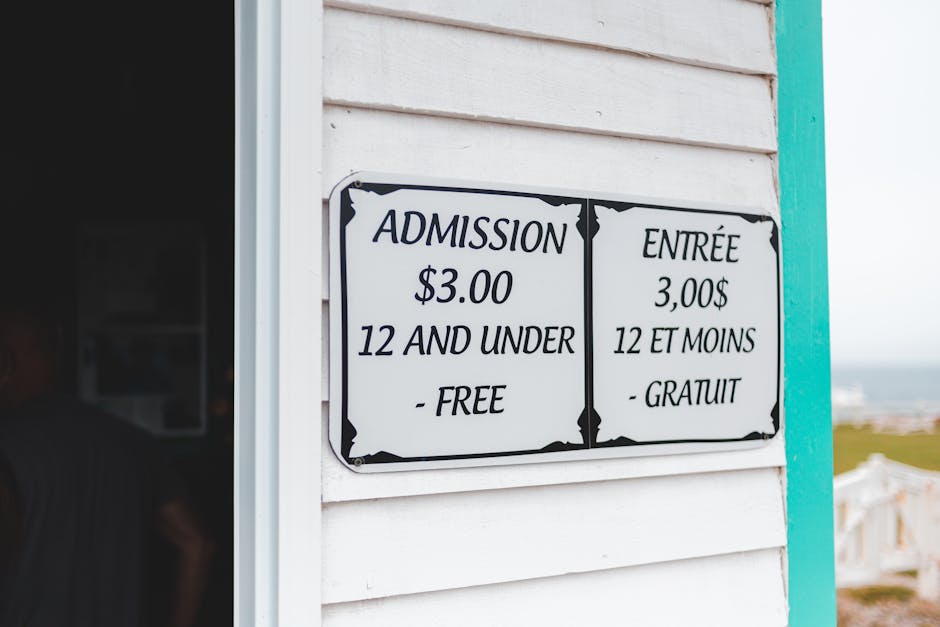 A close-up view of a white wooden building with horizontal siding, featuring a rectangular sign displaying admission prices in English and French. The sign indicates a fee of three dollars for individ
