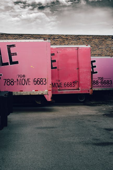A row of three large pink moving containers parked on a paved surface adjacent to a brick building under a cloudy sky. Each container has black lettering indicating a moving company's contact details, with the company's name, Man with Van Yiewsley, associated with the removals service. The containers are positioned closely together, with the leftmost container slightly overlapping the middle one. The containers appear to be made of metal or wood, with some visible hinges and locking mechanisms. No people or other objects are visible in the image. This scene reflects the logistics involved in home relocation, specifically the transportation of furniture and moving boxes using these secure storage units as part of a professional furniture transport and packing process, supporting the content relevance of the page about Yiewsley street-specific move times.