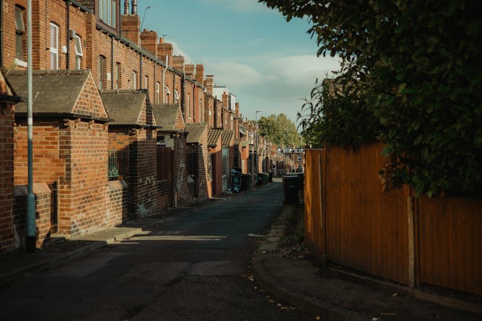 A quiet residential street in Yiewsley with a row of terraced brick houses on the left side, featuring small front yards and tiled roof extensions. The pavement is narrow, with shadows cast by the houses and nearby trees, and a few black wheelie bins are lined up along the kerb. On the right side, a tall wooden fence runs alongside a hedge, partially blocking the view of the property behind it. The street appears to be in preparation for a home relocation, with outdoor space clear of obstructions and ready for the load-in process, reflecting typical conditions suitable for house removals and furniture transport services. The scene is under natural daylight with a partly cloudy sky, emphasizing the typical surroundings where [COMPANY_NAME] would carry out packing, moving, and loading activities as part of their residential removal services.