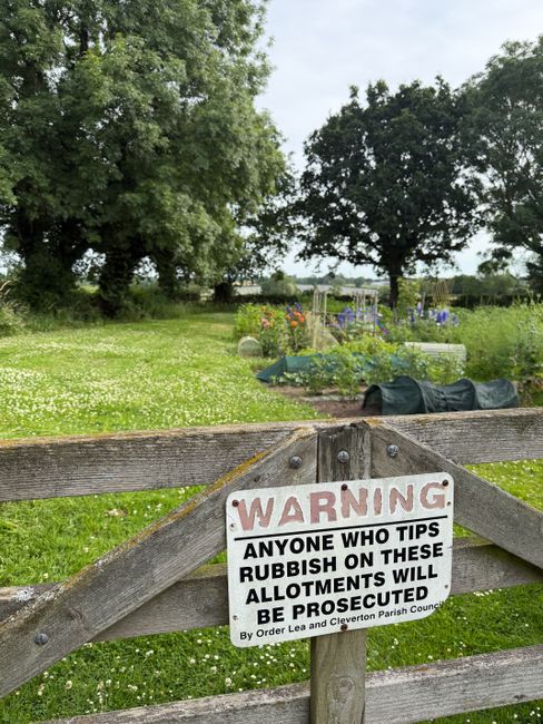 A close-up of a weathered wooden fence with a white warning sign attached, reading 'WARNING: ANYONE WHO TIPS RUBBISH ON THESE ALLOTMENTS WILL BE PROSECUTED by order Lea and Clevendon Parish Council'. Behind the fence, there is a garden or allotment area with green grass, some flowering plants, and gardening equipment such as black fabric-covered compost bins or covers. In the background, large leafy trees and a partly cloudy sky are visible, indicating an outdoor rural or semi-rural setting. This scene suggests a community garden space that is well-maintained and protected, contrasting with the context of house removals and moving services offered by Man with Van Yiewsley, which might involve clearing or managing similar outdoor areas during a property transfer.